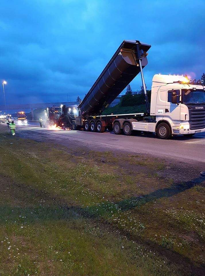 Vörubíll með upplyftan sturtupall losar efni á vegkafla í kvöldrökkri.