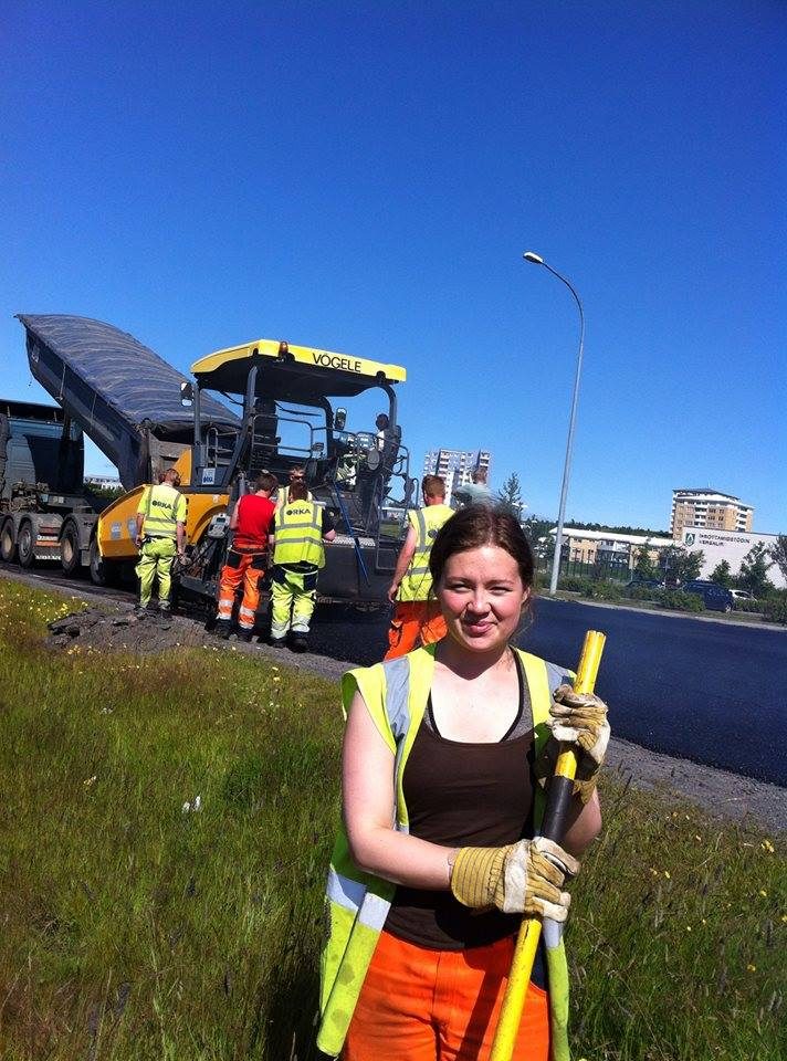 Starfsmaður í endurskinsvesti með skóflu framan við malbikunarvél og vinnuflokk.